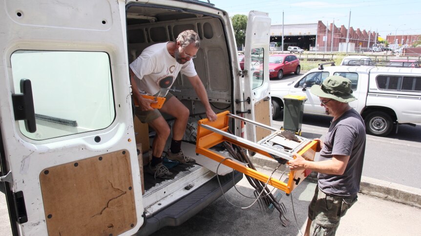 Two men, one in the back of a van, one on the ground, hold a bench saw.