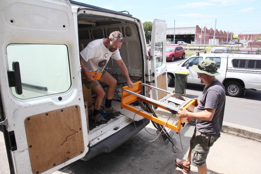 Two men, one in the back of a van, one on the ground, hold a bench saw.