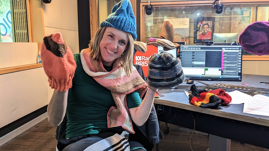 A woman wearing beanies on her head and two hands smiles in a studio surrounded by more beanies