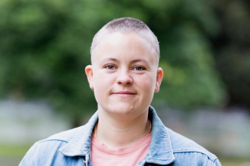 Non-binary person with bald head smiling, wearing pink shirt and denim jacket 