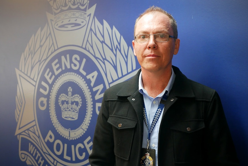 A police officer in plain clothes stands next to the Queensland Police Service logo on a wall
