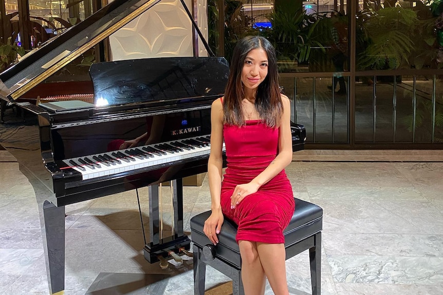 A young women wearing a red dress sits in front of a grand piano.