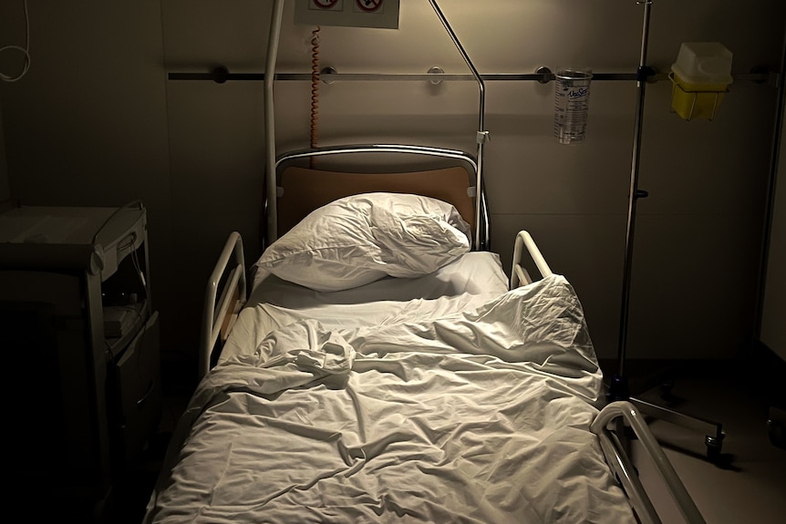 An empty hospital bed in a dark room is lit up by an overhead lamp