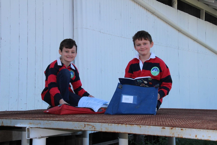 Two boys, aged about 10yo, wearing matching jerseys sit with their school bags and books.