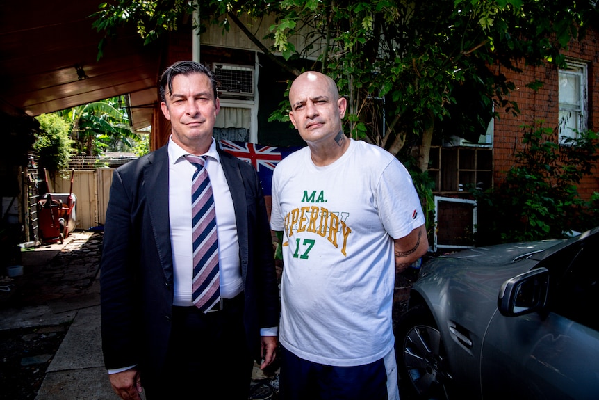 Indigenous man standing next to a man in a suit, outside a home.
