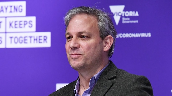 Brett Sutton, dressed in a shirt and jacket, delivers a press conference in front of a purple background.