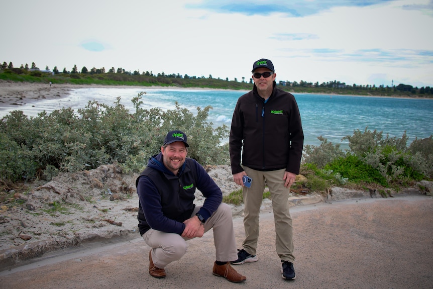 Двое мужчин из IT-компании с черными флисовыми джемперами и кепками стоят на пляжной дорожке