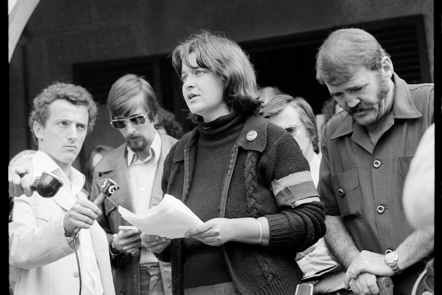 A young woman giving a speech surrounded by people and a fake tombstone.