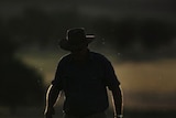 A man with a hat in silouhette, kicking up dust as he walks through a field