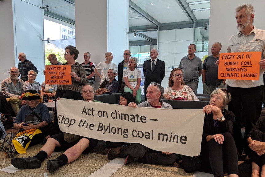 20 people in the foyer of a building hold signs reading 'Act on climate - stop the Bylong coal mine'