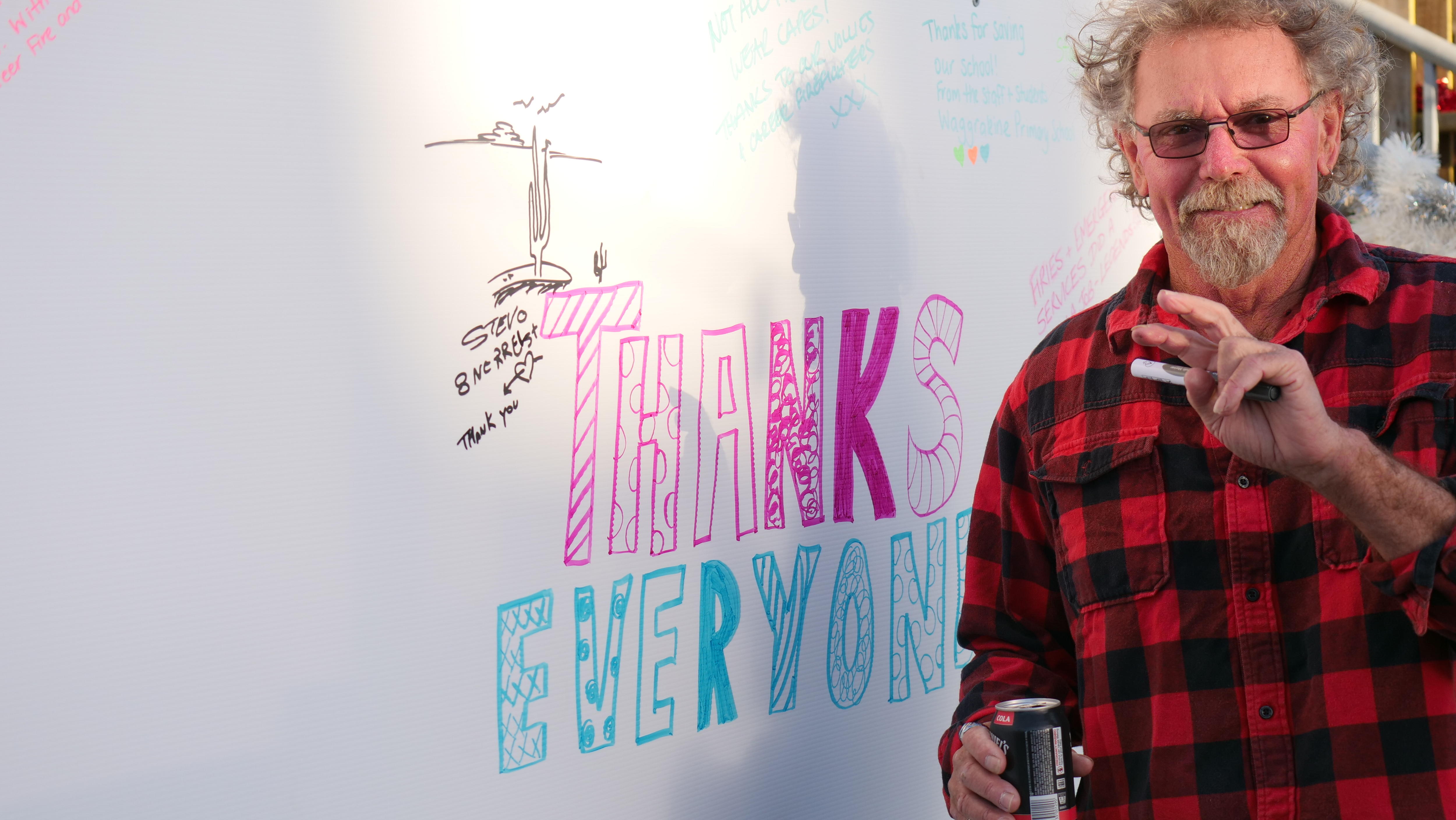 Un hombre con camisa a cuadros y gafas sonríe a la cámara después de escribir en un tablero de agradecimiento. 