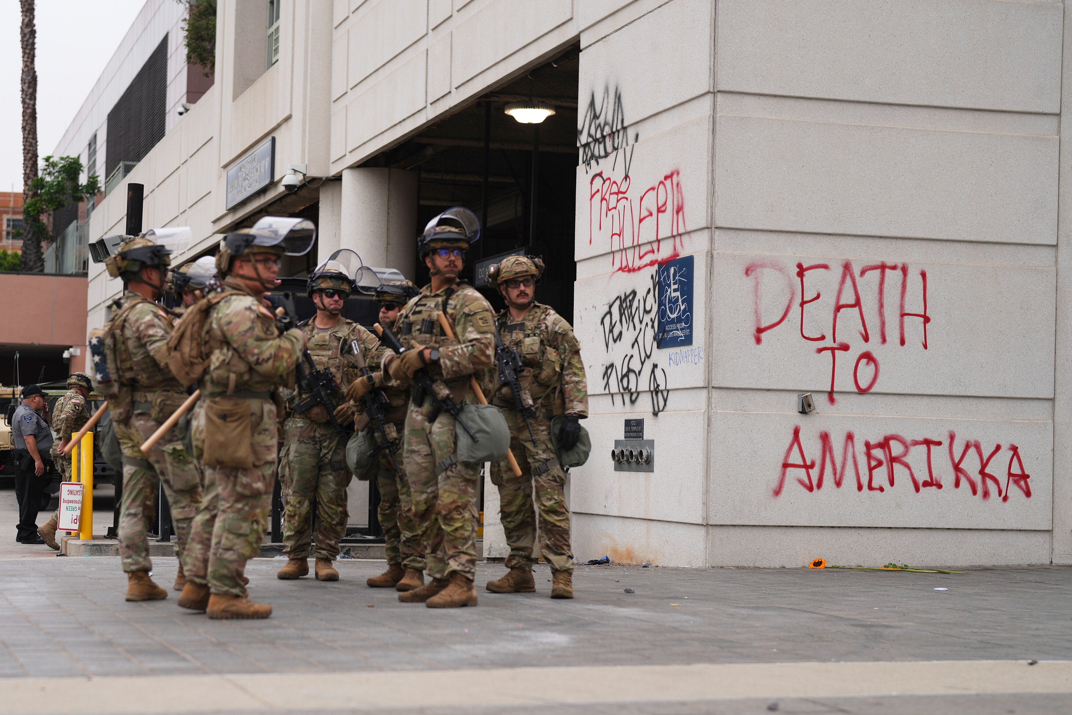 Military members stand next to a wall sprayed with red graffiti 