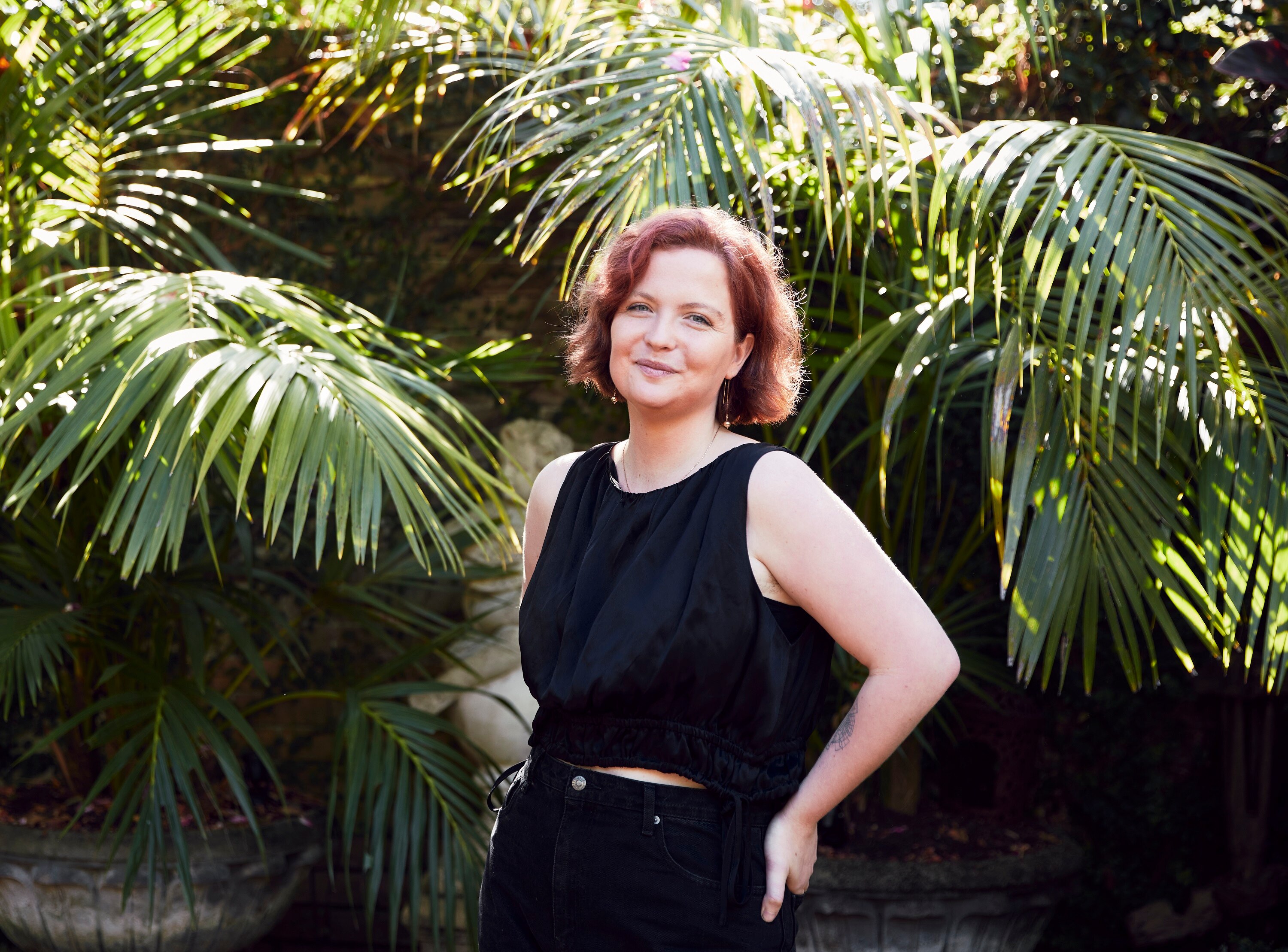 A young white woman with chin-length red hair wearing black stands side on against a backdrop of green leafy plants
