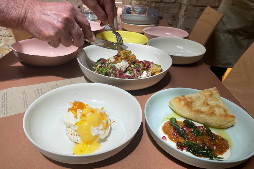 Three plates of food. A salad is being cut by a person holding a knife and fork towards the back of the photo.