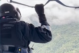 A person in a helmet and protective gear stands at the window of a helicopter, looking out over clouds and forested hills