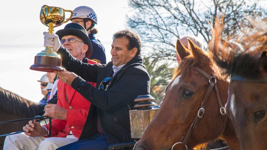 A man sitting on a horse drawn carriage holding up the gold melbourne cup with horse heads in the foreground