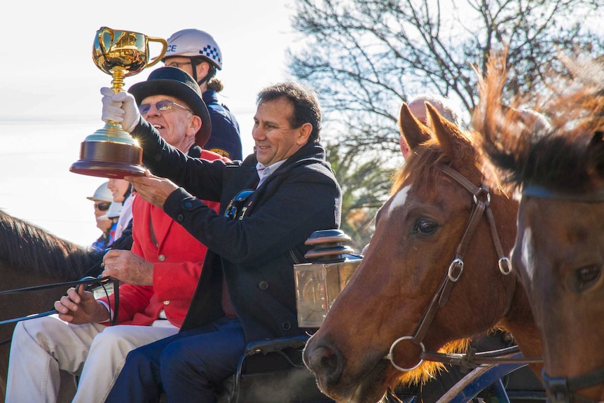 A man sitting on a horse drawn carriage holding up the gold melbourne cup with horse heads in the foreground