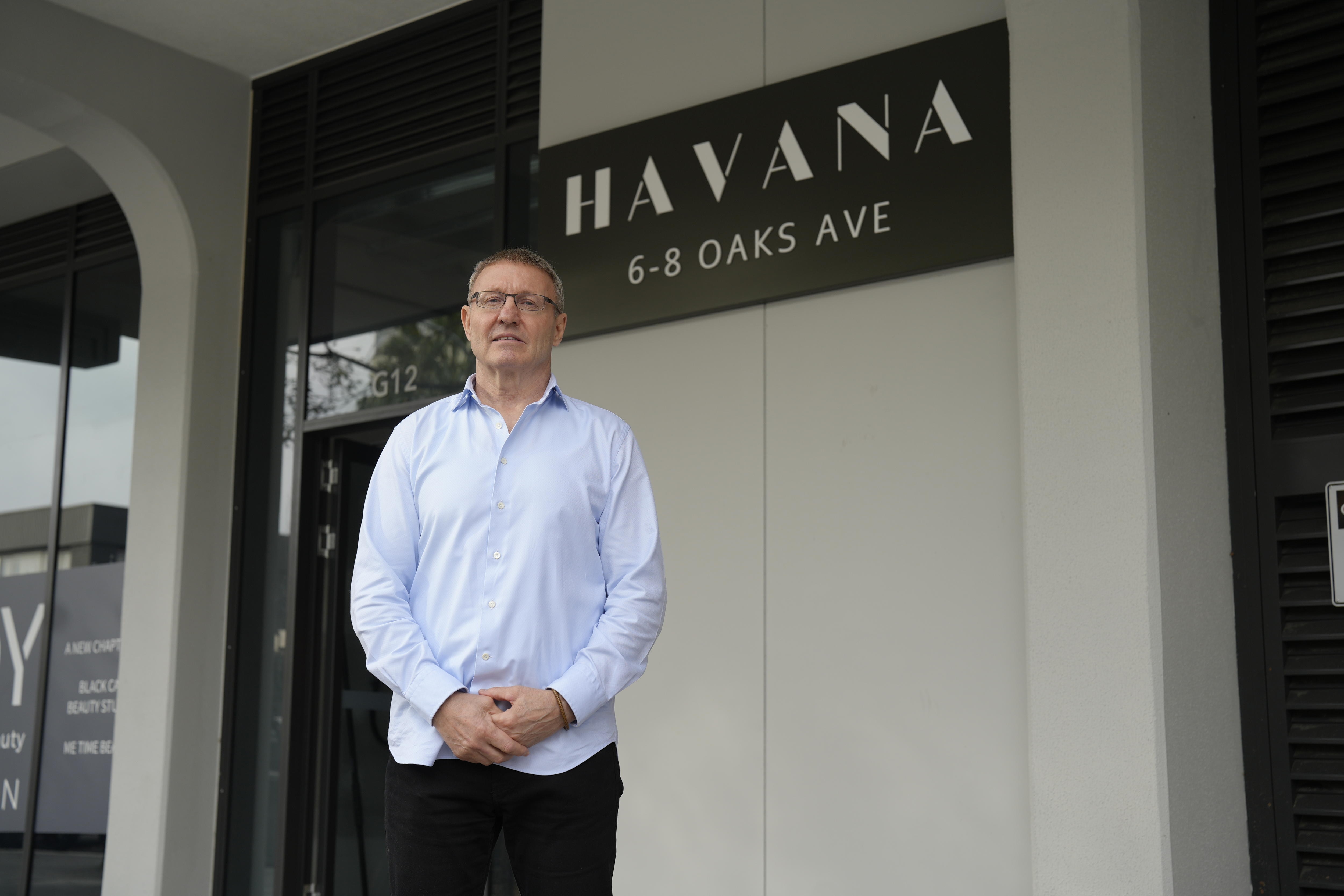 John wears glasses and a shirt, holding his hands in front. He is in front of a building with a sign reading 'Havana'.