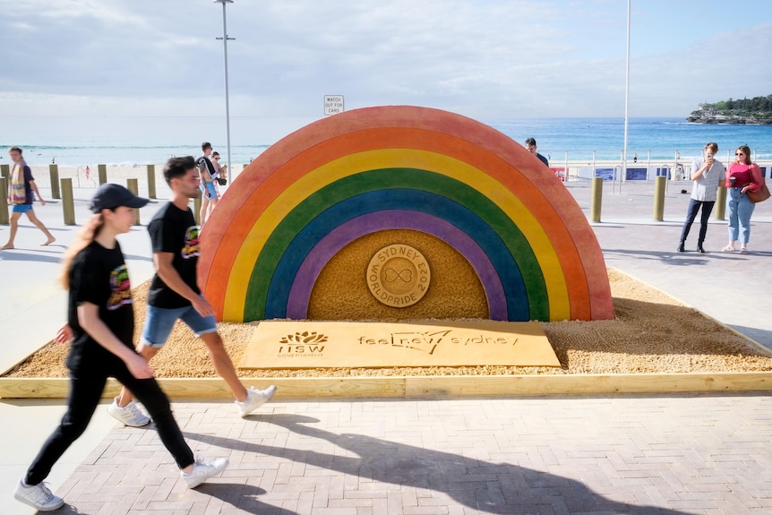 Two people walking past a large rainbow sand sculpture at Bondi Beach