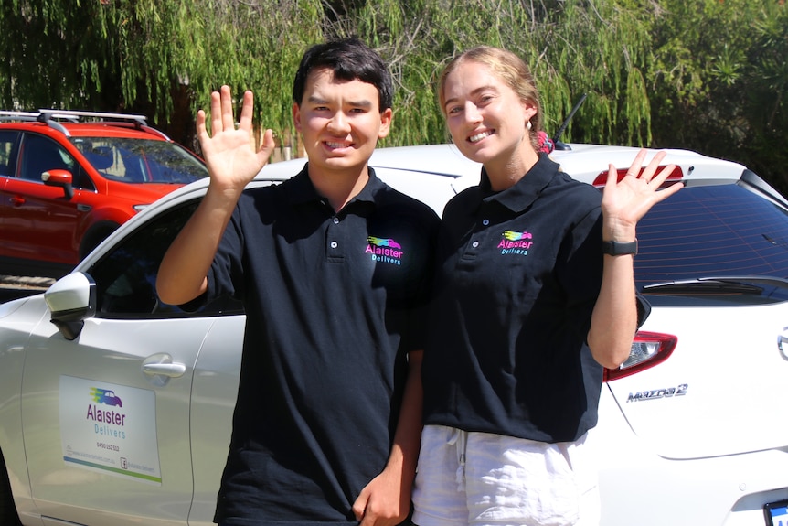 A young man and woman smile and wave standing in front of a white car