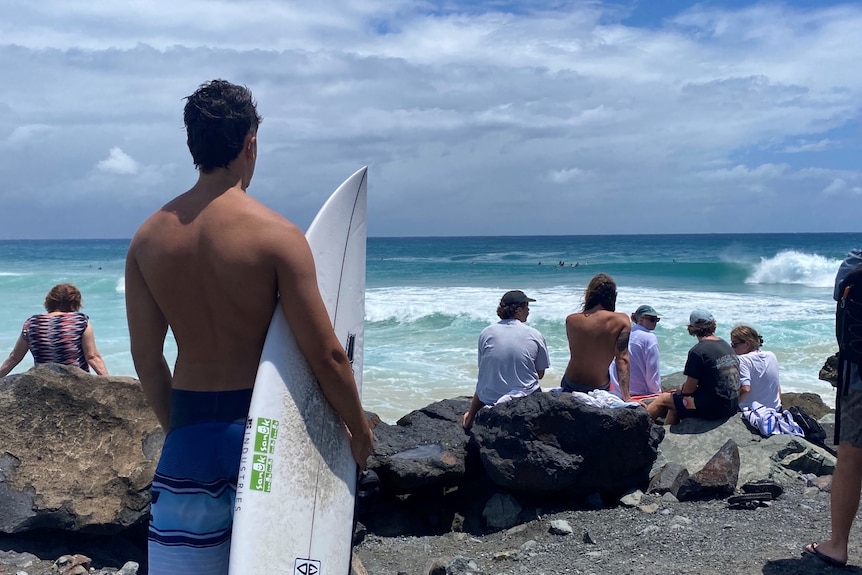 A man stands with a surfboard looking at the waves, people also sit on rocks looking at waves.