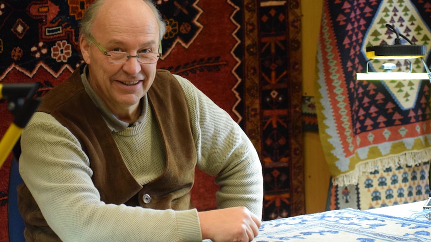 A man smiling at the camera, he is wearing a cream coloured wool jumper and brown suede vest