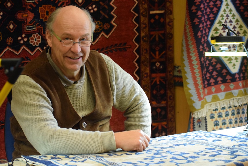 An older, bespectacled man sits with his arms resting on a carpet he is working on, smiling.