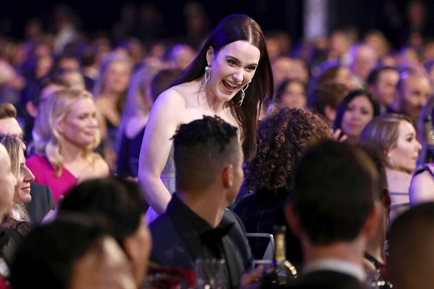 Rachel Brosnahan grins as she walks through the crowd to the stage to accept award wearing drop earrings and silver dress.