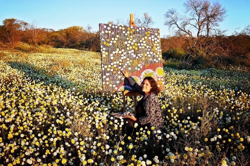 Woman outside painting yellow and white flowers on canvas, blue sky, brown shrubs