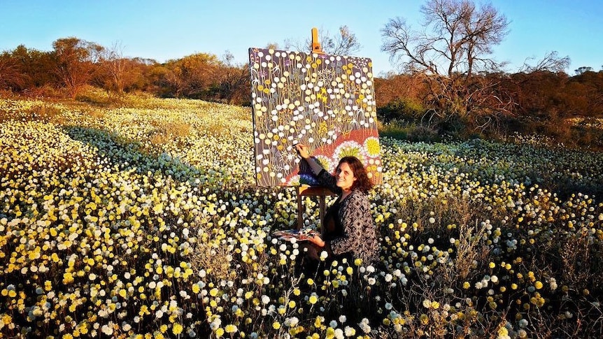 woman outside painting yellow and white flowers on canvas, blue sky, brown shrubs