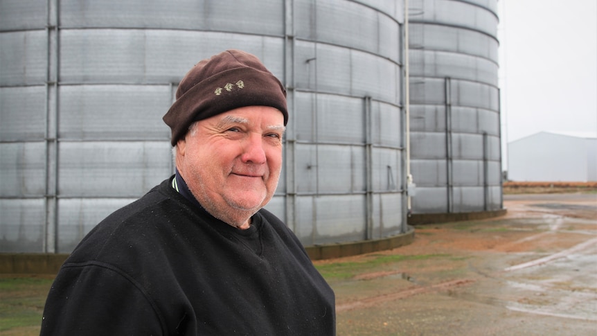 A man wearing a beanie stands in front of two silos.