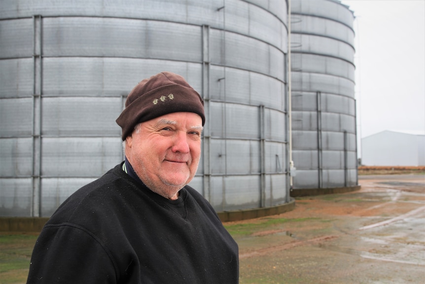 A man wearing a beanie stands in front of two silos.