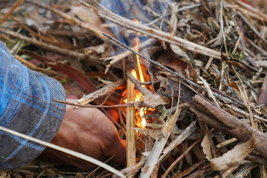 A hand lights a small orange flame among dry twigs.