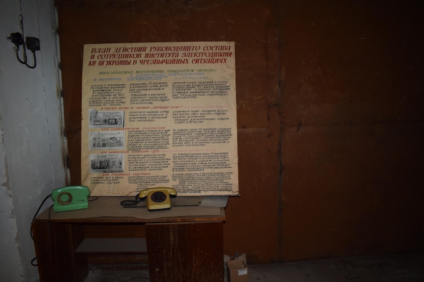 Two rotary telephones sit on a dusty desk in a dark room. A sign with text written in Ukrainian is propped up against the wall