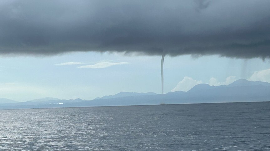 Long range shot of a water spout appearing over the ocean with land behind it.
