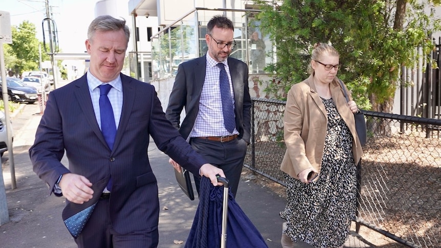 A blonde, bespectacled woman and two men in suits walk down a city street.