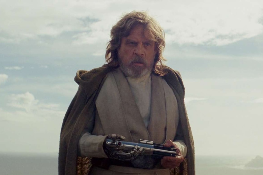 Luke Skywalker in a cloak looking perturbed and holding his old lightsaber in his hands.