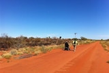 A man in a wheelchair and another man ride and walk on a red dirt road in outback Northern Territory.