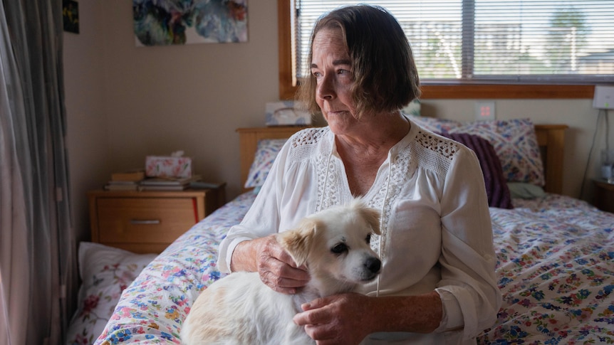 A woman sits on her bed with a dog on her lap.