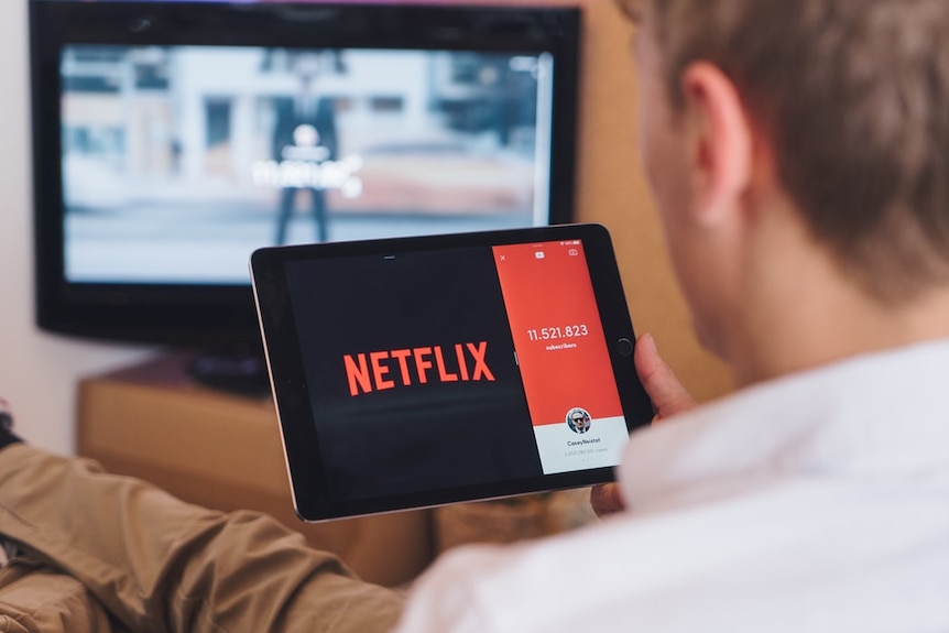 A man with dark blonde hair watches Netflix on a tablet, with a TV on in the background.