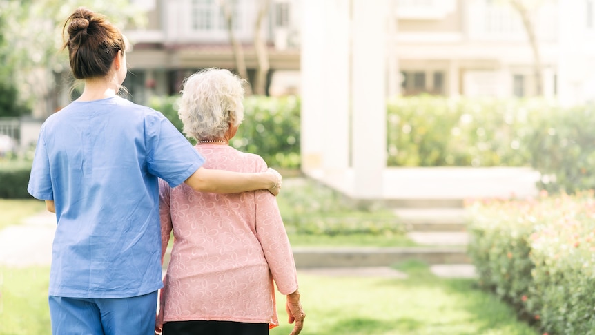 A woman wearing blue scrubs has her arm around an older woman wearing a pink cardigan as they walk across a lush green lawn.