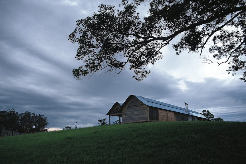 A wooden and corrugated iron house sitting on a grassy hill with clouds in the sky.