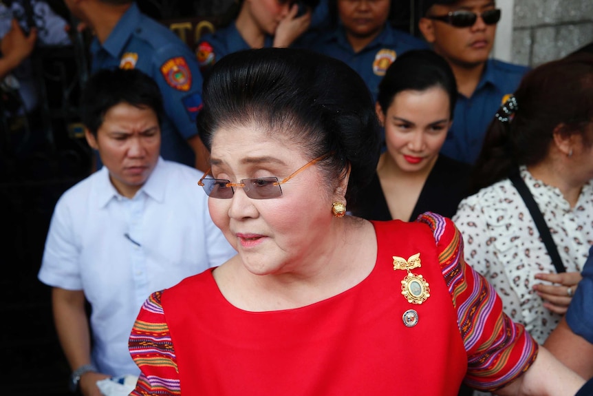 A woman with a bouffant, glasses and a bright red top surrounded by police and other people.