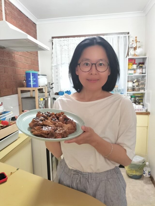 An smiling, bespectacled Chinese woman in a kitchen holds a plate of succulent chicken towards the camera.