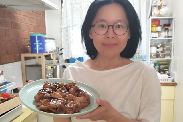 An smiling, bespectacled Chinese woman in a kitchen holds a plate of succulent chicken towards the camera.