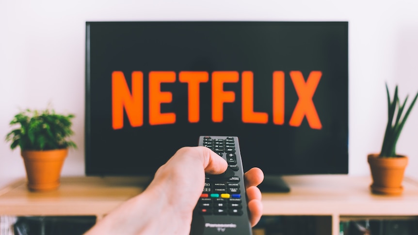 A hand pointing a TV remote towards a TV, which displays the Netflix logo