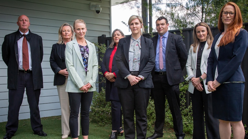 Eight people stand outside a house and fence.