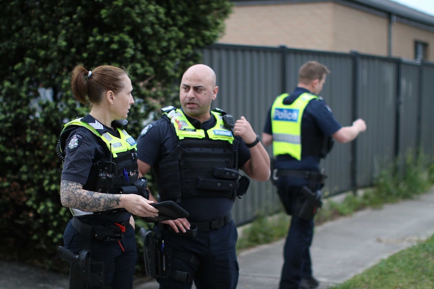 Three police officers stand in a suburban street, outside a property with a green fence.