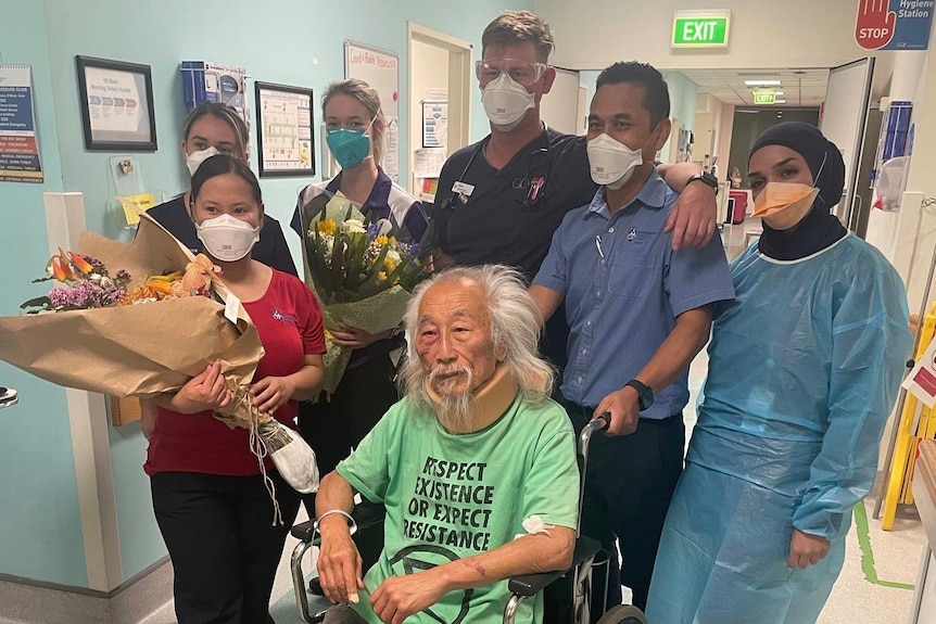 An elderly man wearing a neckbrace sits in a wheelchair with medical staff around him