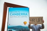 A man holds a sign reading "pay up" in front of a sign saying "Welcome to Canberra, Australia's capital".